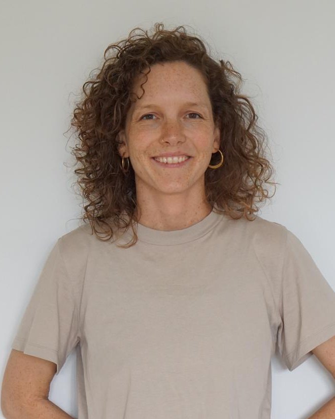 Porträt von Annica Bauer: lächelnde Frau mit braunen Locken und goldenen Creolen vor einer weißen Wand.
