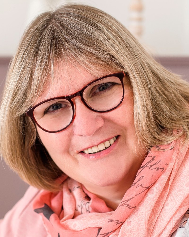 Porträt von Edith Sauerbier: lächelnde Frau mit roter Brille, blondem Bob und rosa Schal.